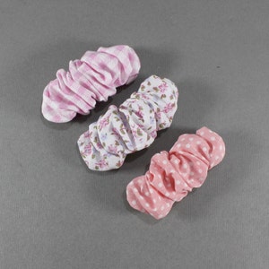 May include: Three fabric scrunchies are displayed. One features a pink and white gingham pattern, another has a floral design on white, and the third is pink with white polka dots. The scrunchies are gathered and likely made of cotton.