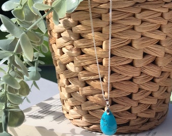 Puede incluir: Un collar de cadena de plata con un colgante de lágrima turquesa. El collar está colgado de una cesta de mimbre.