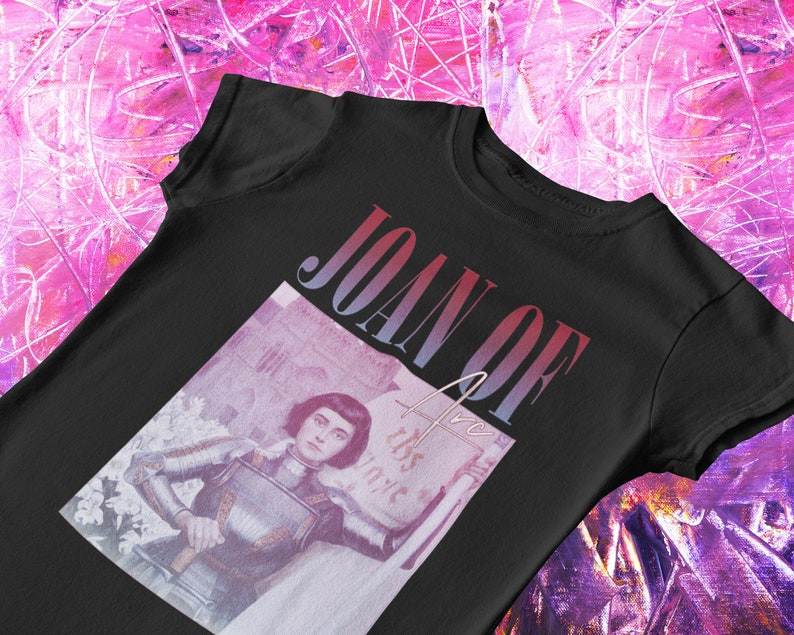 May include: Black t-shirt with a vintage-style image of Joan of Arc in armor, holding a sword and a banner that reads "This is my voice". The text "JOAN OF ARC" is printed in a gradient font across the top of the image.