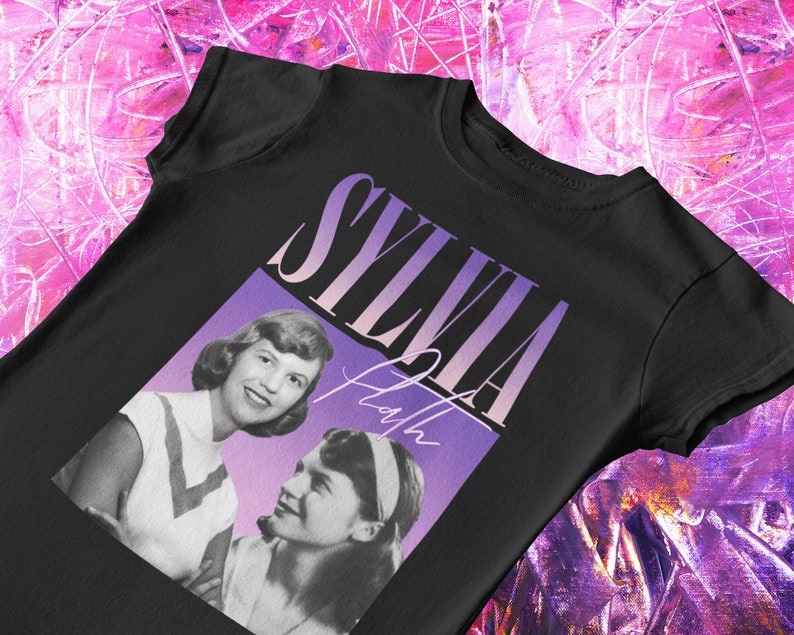 May include: Black t-shirt with a graphic of Sylvia Plath and the text "Sylvia Plath" in a purple and pink gradient.