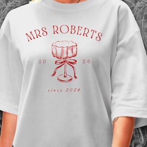 May include: White t-shirt featuring the text "MRS ROBERTS" in red, above a red illustration of a stemmed glass with a bow. The numbers "20 26" and "since 2026" are also in red.