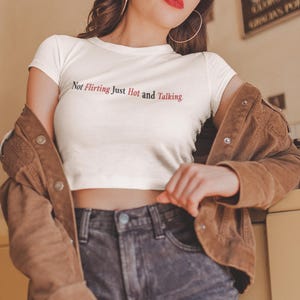 Puede incluir: Un top corto blanco con el texto "Not Flirting Just Hot and Talking" en negro y rojo. Se combina con vaqueros oscuros y una chaqueta de pana marr&oacute;n.