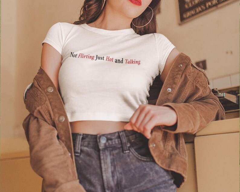 Puede incluir: Un top corto blanco con el texto "Not Flirting Just Hot and Talking" en negro y rojo. Se combina con vaqueros oscuros y una chaqueta de pana marr&oacute;n.