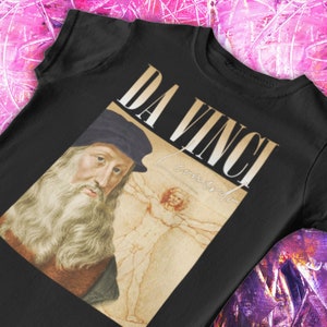 Pode incluir: Camiseta preta com um gráfico de Leonardo da Vinci e o texto "Da Vinci" em uma fonte branca estilizada.