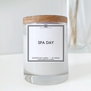 May include: A white soy candle with a wooden lid and a label that reads "SPA DAY" in black text. The label also includes the text "SCENTED SOY CANDLE | 4% AROMA" and "HAND-POURED IN CANADA".
