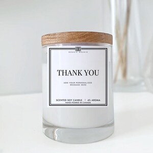 May include: A white soy candle with a wooden lid and a black and white label that says "Thank You" and "Bougies Bougie". The label also includes the text "Add your personalized message here", "Scented Soy Candle | 4% Aroma", and "Hand-poured in Canada".