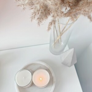 May include: Two white candles in glass jars on a white marble coaster. The candles are lit and the flames are visible. The coaster is on a white surface.