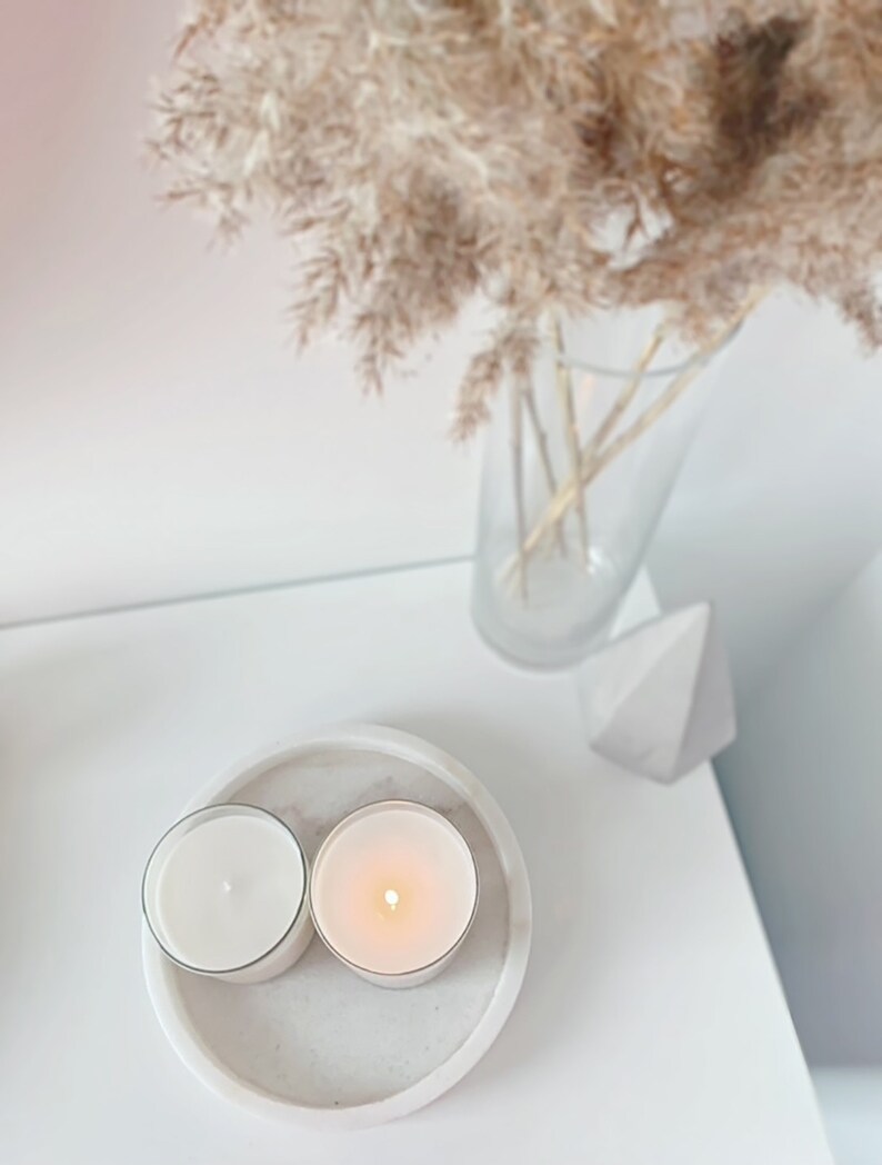 May include: Two white candles in glass jars on a white marble coaster. The candles are lit and the flames are visible. The coaster is on a white surface.