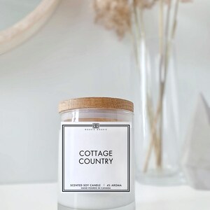 May include: A white soy candle with a wooden lid and a black label that reads "Bougie Bougie Cottage Country Scented Soy Candle | 4% Aroma Hand-Poured in Canada"