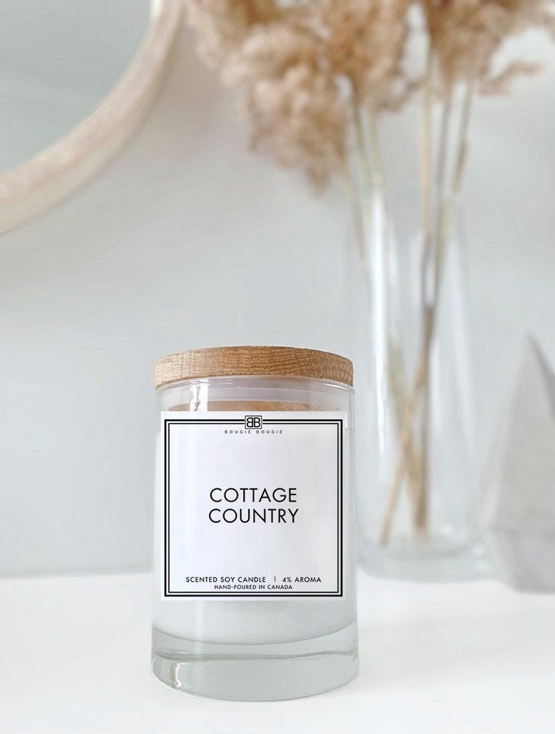May include: A white soy candle with a wooden lid and a black label that reads "Bougie Bougie Cottage Country Scented Soy Candle | 4% Aroma Hand-Poured in Canada"