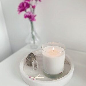 May include: A white candle with a lit wick in a clear glass jar on a white marble tray with two matches and two clear quartz crystals.