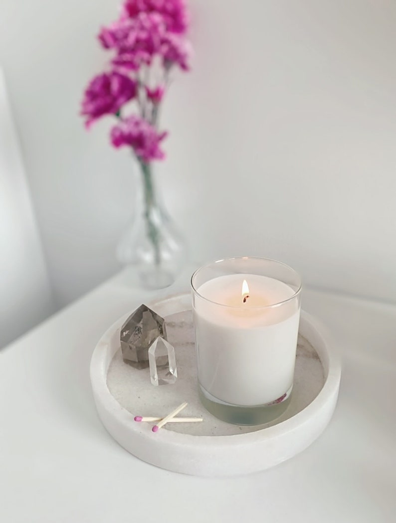 May include: A white candle with a lit wick in a clear glass jar on a white marble tray with two matches and two clear quartz crystals.