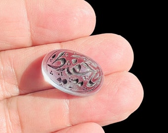 May include: An oval-shaped, silver-colored, carved stone with intricate Arabic calligraphy. The stone is set against a pink background.