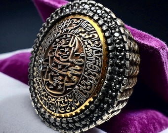 May include: A detailed, ornate ring with a central design featuring Arabic calligraphy. The ring has a silver-colored band with a gold-colored inner circle and intricate detailing. The ring is displayed on a purple velvet surface.
