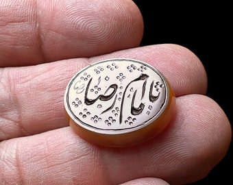 May include: An oval-shaped ring with an engraved design and Arabic script. The ring has a light brown stone and a silver-colored metal band. The ring is held in a person's hand against a black background.
