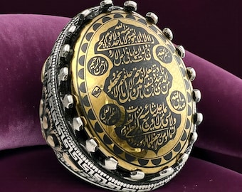 May include: A silver ring with an oval, gold-colored center featuring black calligraphy. The ring has a detailed design around the band and the center stone. The ring is set against a dark purple background.