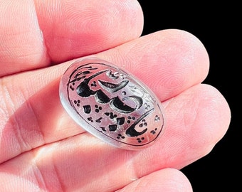 May include: A clear, oval-shaped stone with black calligraphy etched on its surface. The design features flowing script and small dot patterns. The stone is held in a hand against a black background.
