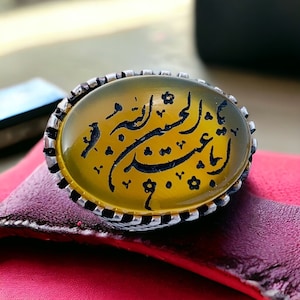 May include: A silver ring with a yellow agate stone set in the center. The stone has black Arabic calligraphy etched into it.