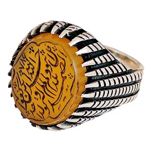 May include: A silver ring with a yellow agate stone set in the center. The stone has Arabic calligraphy etched into it. The band of the ring is decorated with a textured design.
