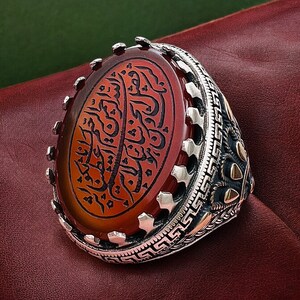 May include: A silver ring with an oval-shaped carnelian stone set in a decorative silver frame. The stone has intricate Arabic calligraphy etched into it.