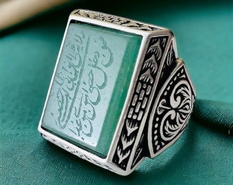 May include: A silver ring featuring an intricate design and a green stone set in the centre. The stone has Arabic calligraphy etched into it.
