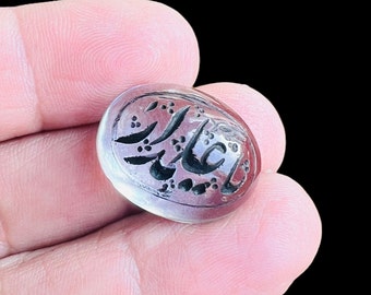 May include: A round, dark red gemstone with intricate black Arabic calligraphy carved into the surface. The gemstone is set in a silver ring.
