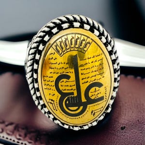 May include: A silver ring with a gold oval inlay featuring Arabic calligraphy and a black inscription. The ring has a braided design around the edge.