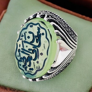 May include: A silver ring with a green stone featuring Arabic calligraphy. The ring has a detailed, braided band.