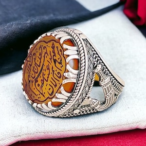 May include: A silver ring with an intricate design featuring a large, round, orange stone with Arabic calligraphy. The ring has a detailed, ornate design with a raised, textured band.
