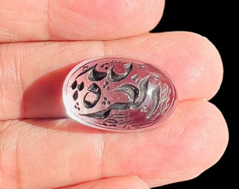 May include: A clear oval-shaped stone with a pink hue and intricate silver calligraphy etched into the surface.