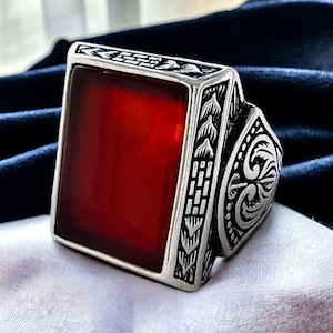 May include: A silver ring with a large, rectangular, red gemstone set in the center. The ring has intricate, detailed designs on the band.