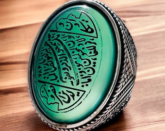 May include: A silver ring featuring an oval, light green stone with black calligraphy. The ring has a detailed, textured band. The ring is set on a wooden surface.
