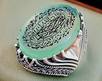 May include: A silver ring with a green stone featuring Arabic calligraphy. The ring has an intricate design with a braided band.
