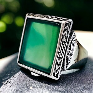 May include: A silver ring with a square green stone set in a detailed silver frame. The ring has an intricate design on the band.