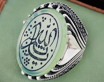 May include: A silver ring with a green stone featuring Arabic calligraphy. The stone is oval shaped and set in a silver band with intricate designs.