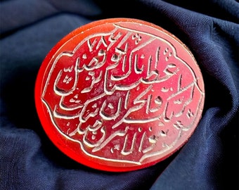 May include: A red carnelian stone with intricate Arabic calligraphy carved into the surface. The stone is round and has a smooth, polished finish.