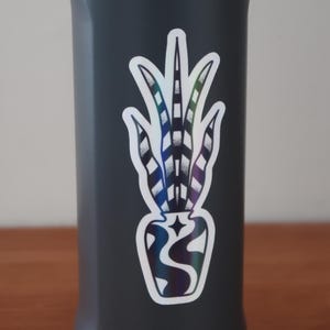 Peut inclure: Une gourde gris foncé avec un autocollant holographique d'une plante dans un vase. L'autocollant a un contour blanc et présente un design détaillé avec des nuances de bleu, de violet et de vert.