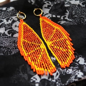 May include: A pair of long, beaded earrings in a teardrop shape. The earrings are made with orange and yellow beads and have a gold-toned metal hook.