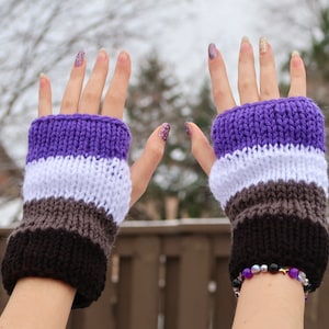 May include: A pair of knitted fingerless gloves with purple, white, grey, and black stripes. The gloves are being worn by a person with their hands raised.