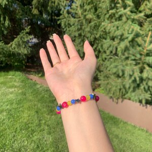 Pansexual pride flag gemstone bracelet being worn on someone's wrist. There are green bushes in the background and it is sunny.