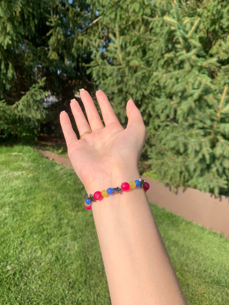 Pansexual pride flag gemstone bracelet being worn on someone's wrist. There are green bushes in the background and it is sunny.