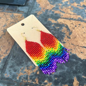 May include: A pair of beaded earrings in a rainbow gradient, from red to purple. The earrings are attached to gold-colored hooks and are displayed on a tan card.