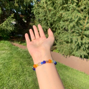 Aroace pride flag gemstone bracelet being worn on someone's wrist. There are green bushes in the background and it is sunny.