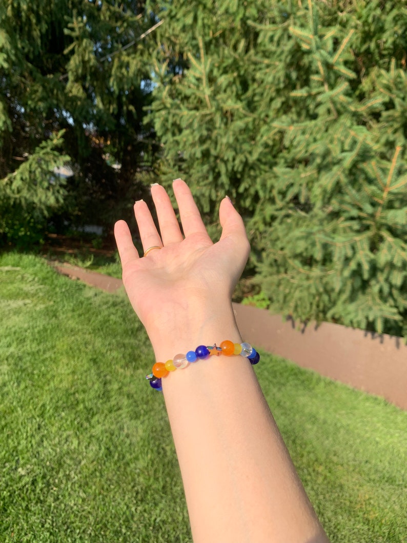 Aroace pride flag gemstone bracelet being worn on someone's wrist. There are green bushes in the background and it is sunny.