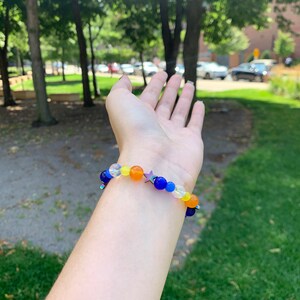 Aroace pride flag gemstone bracelet being worn on someone's wrist. There are green bushes in the background and it is sunny.