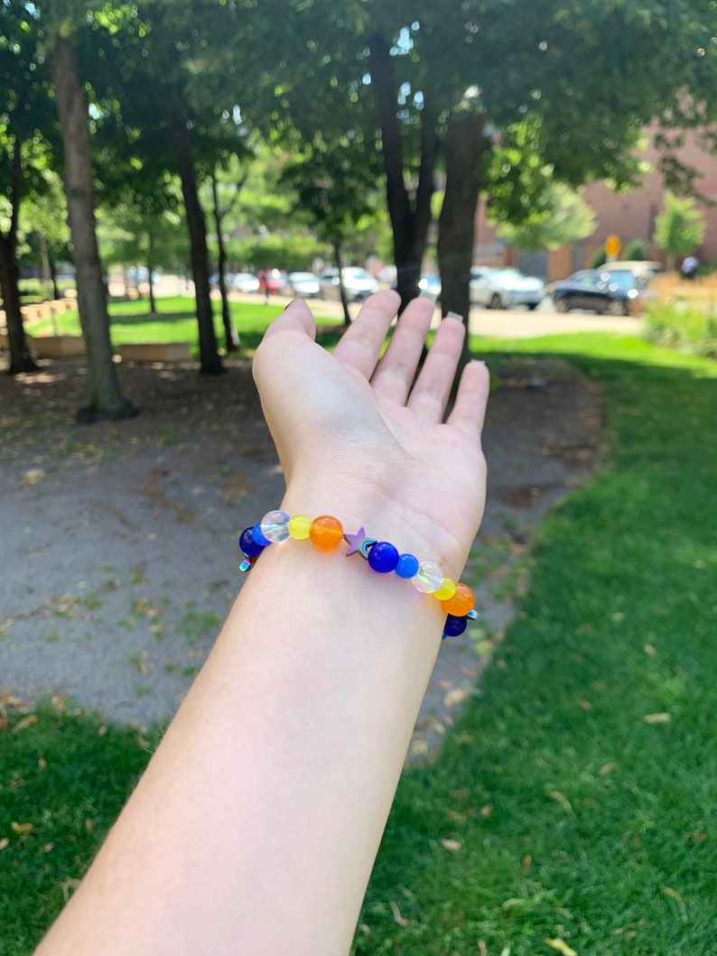 Aroace pride flag gemstone bracelet being worn on someone's wrist. There are green bushes in the background and it is sunny.