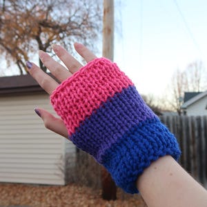 以下が含まれることがあります： ピンク、紫、青のストライプのニットリストウォーマー。リストウォーマーは手に着用されています。背景には家、木、曇り空が写っています。