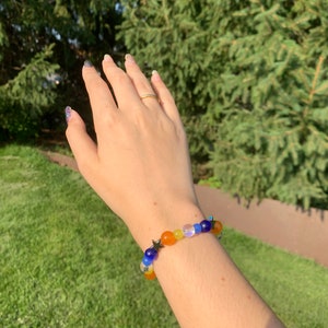 Aroace pride flag gemstone bracelet being worn on someone's wrist. There are green bushes in the background and it is sunny.