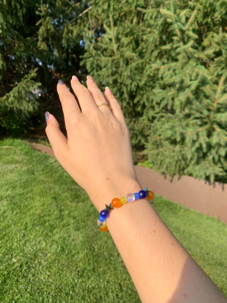 Aroace pride flag gemstone bracelet being worn on someone's wrist. There are green bushes in the background and it is sunny.