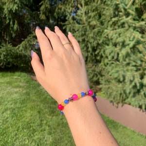 Pansexual pride flag gemstone bracelet being worn on someone's wrist. There are green bushes in the background and it is sunny.